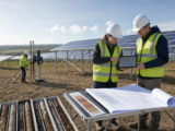 Two engineers review a structural model and controlled site drawings beside soil samples, a trial installation rig and ground-mounted photovoltaic rows.
