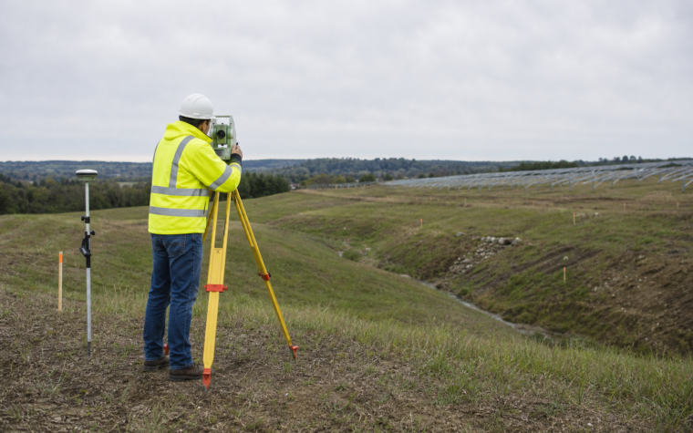 A surveyor uses a total station on rolling terrain beside a visible swale, with ground-mount solar support rows in the distance.